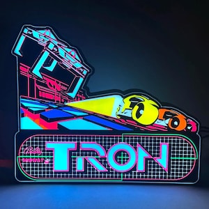 May include: A glowing LED sign featuring the TRON arcade game logo in bright cyan, pink, and yellow. The design includes a stylised vehicle graphic, a grid pattern background, and the Bally Midway text, all illuminated against a dark backdrop.