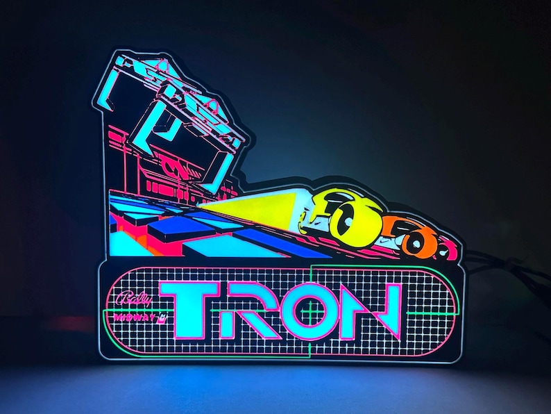 May include: A glowing LED sign featuring the TRON arcade game logo in bright cyan, pink, and yellow. The design includes a stylised vehicle graphic, a grid pattern background, and the Bally Midway text, all illuminated against a dark backdrop.