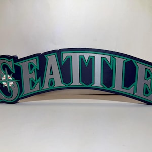 May include: A curved wall sign featuring the word SEATTLE in large, block-style silver letters with dark blue and teal outlines. The letter S is replaced by a compass rose graphic with a baseball design at its center, rendered in white, red, and teal.