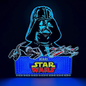 May include: A neon sign featuring Darth Vader's silhouette, a TIE fighter, and an X-wing fighter. The sign has the Atari Star Wars logo in yellow and red, with a blue grid background. The sign is illuminated with blue light.