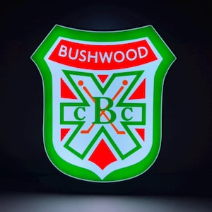 May include: A shield-shaped illuminated sign featuring the text BUSHWOOD in white on a red background. The center displays a large green X with two crossed golf clubs and the letters C, B, and C in green, set against a white and red background.