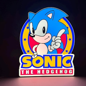 May include: A glowing LED sign featuring the character Sonic the Hedgehog. The design includes a blue hedgehog character in front of a yellow ring with blue stars, with the text SONIC THE HEDGEHOG displayed in bold yellow and white lettering.