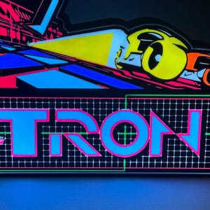 May include: A rectangular LED sign featuring the word TRON in large blue letters with pink outlines. Below the text, a grid pattern is displayed. Above, a yellow and orange vehicle graphic moves across a dark surface with blue and pink geometric shapes.