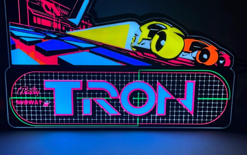 May include: A rectangular LED sign featuring the word TRON in large blue letters with pink outlines. Below the text, a grid pattern is displayed. Above, a yellow and orange vehicle graphic moves across a dark surface with blue and pink geometric shapes.