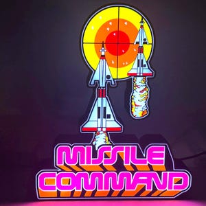 May include: A light-up sign featuring the text Missile Command in bright pink and orange letters. Above the text are two white and red rockets launching toward a yellow and orange circular target graphic with a black crosshair overlay.