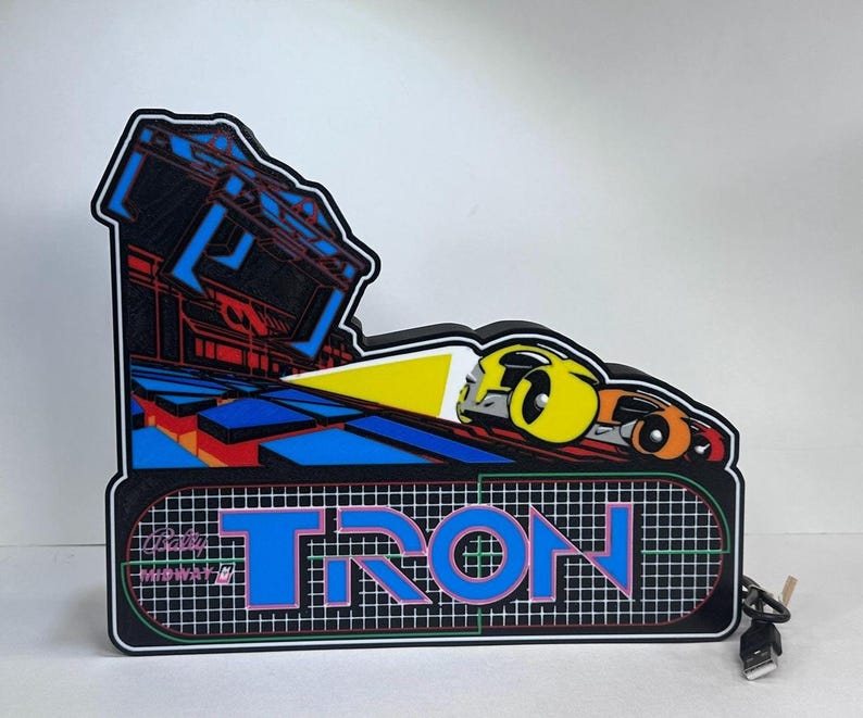 May include: A decorative Tron arcade sign featuring a black background with a grid pattern, the blue Tron logo, and a graphic of a yellow and orange light cycle. The sign includes a black power cord with a USB connector extending from the right side.