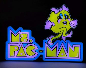 May include: A glowing LED sign featuring the character Ms. Pac-Man in yellow with a pink bow and high heels, positioned above the text Ms. Pac-Man in bold yellow letters with a pink outline, all set against a vibrant blue illuminated background.