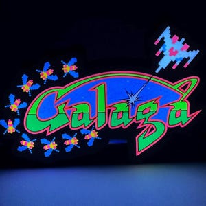 May include: A neon sign with the word "Galaga" in green and pink, with a blue background. The sign features pixelated space ships and alien insects, evoking a retro arcade game theme.