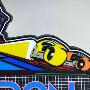 May include: A multi-coloured plastic sign featuring a stylised yellow and orange car graphic with a bright yellow headlight beam. The design includes a black background with a white grid pattern and partial blue and red geometric shapes above.