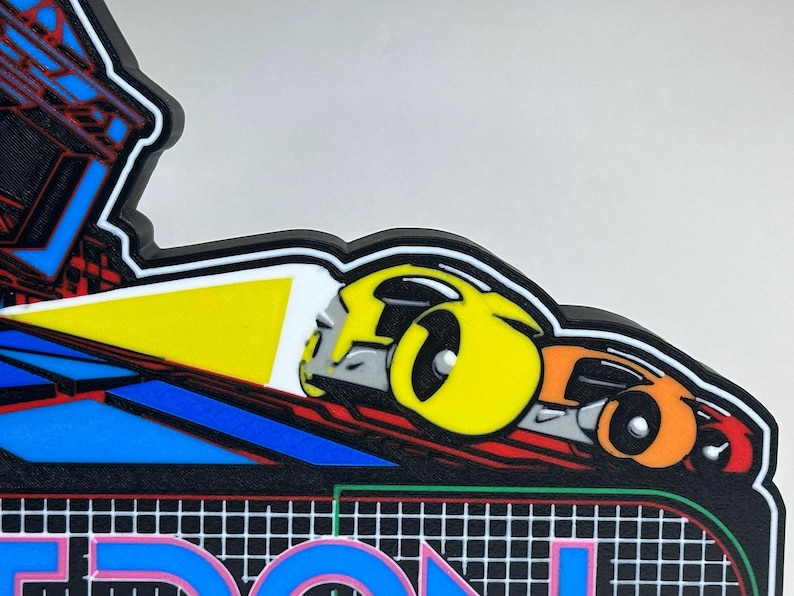 May include: A multi-coloured plastic sign featuring a stylised yellow and orange car graphic with a bright yellow headlight beam. The design includes a black background with a white grid pattern and partial blue and red geometric shapes above.