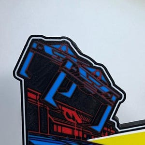 May include: A die-cut sticker featuring a stylised graphic of a dark structure with vibrant blue and red geometric line art. The design has a thick black border and a white outline, showcasing a sharp, angular aesthetic with high-contrast colours.