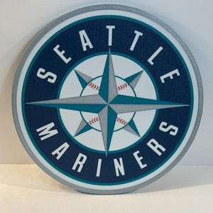 May include: Circular Seattle Mariners logo with a navy blue background and a silver border. The logo features a compass rose design in white, teal, and silver, with baseball stitching accents. The words "SEATTLE MARINERS" are in white, arched around the compass rose.