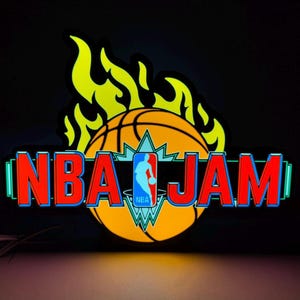 May include: An illuminated NBA JAM sign featuring a large orange basketball with yellow flames rising from the top. The text NBA JAM is displayed in bold red letters with blue outlines, centered over the basketball with the blue NBA logo silhouette.