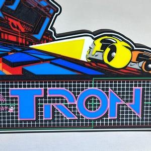 May include: A decorative wall sign featuring the Tron arcade game logo with the text Bally Midway and TRON in blue letters over a white grid. The design includes a yellow and orange light cycle graphic on a black background with blue geometric shapes.