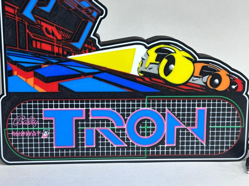 May include: A decorative wall sign featuring the Tron arcade game logo with the text Bally Midway and TRON in blue letters over a white grid. The design includes a yellow and orange light cycle graphic on a black background with blue geometric shapes.