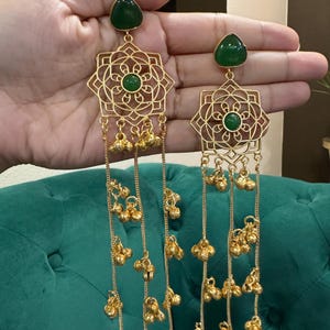 May include: Gold earrings with a geometric pattern and dangling bell charms. Each earring has a green gemstone at the top and centre. The earrings are shown against a green backdrop.