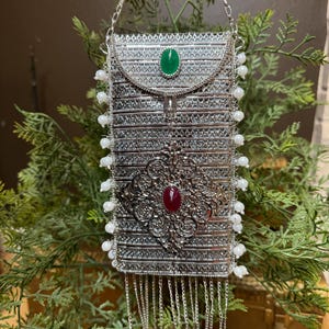 May include: A silver-toned, rectangular handbag with intricate metalwork and a chain strap. It features a green oval stone on the flap and a red oval stone in the center. White bead accents line the sides, and silver fringe hangs from the bottom.