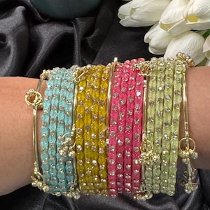 May include: A collection of colorful bangles in shades of blue, yellow, pink, and green. Each bangle is adorned with small, sparkling embellishments and gold accents. The bangles are displayed on a black surface, with a candle and white flowers in the background.