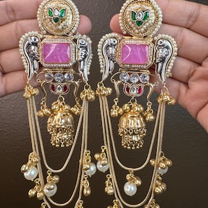 May include: A pair of ornate gold-toned earrings with a pink gemstone center. The earrings feature intricate detailing, including pearls, silver accents, and dangling chains with pearl and gold bead clusters. The top has a green and gold design.