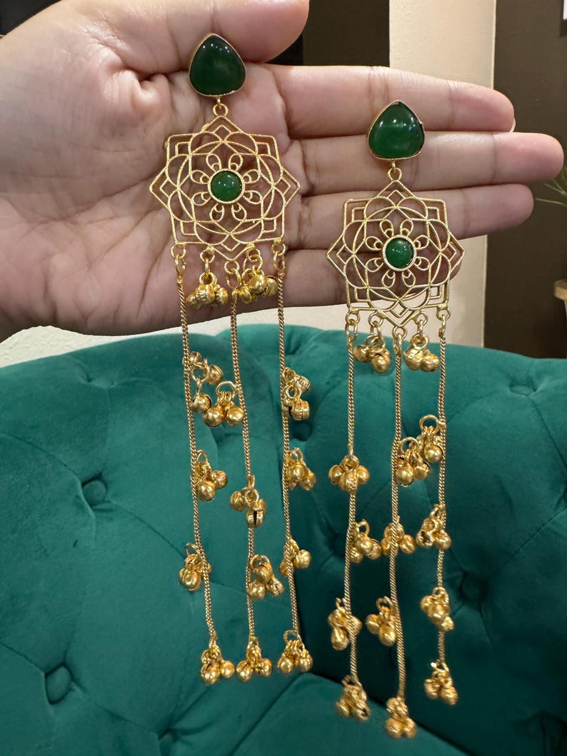 May include: Gold earrings with a geometric pattern and dangling bell charms. Each earring has a green gemstone at the top and centre. The earrings are shown against a green backdrop.