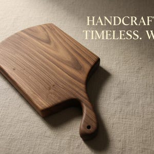 May include: A brown wooden cutting board with a handle and a hole for hanging, featuring a visible natural grain pattern. The text HANDCRAFTED. TIMELESS. WOOD. is printed in the upper right corner of the image in a light beige sans-serif font.