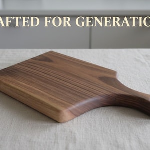 May include: A rectangular wooden cutting board with a handle featuring a circular hole at the end. The board displays a rich brown wood grain pattern. The text CRAFTED FOR GENERATIONS is printed in a light cream color above the wooden board.