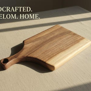 May include: A rectangular wooden cutting board with a handle featuring a circular hole. The wood displays natural grain patterns in shades of light and dark brown. The text HANDCRAFTED. HEIRELOM. HOME. is printed in the upper left corner of the image.