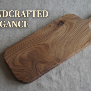 May include: A rectangular wooden serving board with a handle featuring a hole for hanging. The wood displays a prominent natural grain pattern in shades of light and dark brown. The text HANDCRAFTED ELEGANCE is printed in the upper left corner.
