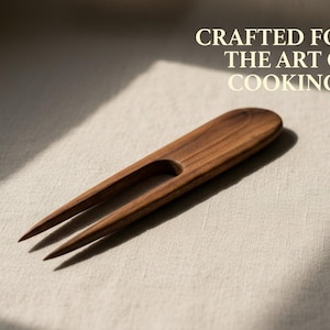 Può includere: Una forchetta da cucina in legno con due rebbi e un manico. La forchetta è in legno marrone scuro ed è posta su uno sfondo neutro. Il testo in alto recita "Crafted for the Art of Cooking."