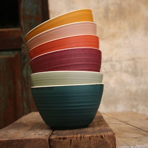 Könnte beinhalten: Ein Stapel von sechs bunten Keramikschalen. Die Schalen sind in den Farbtönen Gelb, Rosa, Orange, Burgunderrot, Hellgrün und Türkis gehalten. Jede Schale hat eine strukturierte Oberfläche und einen weißen Rand. Sie sind auf einer rustikalen Holzoberfläche gestapelt.