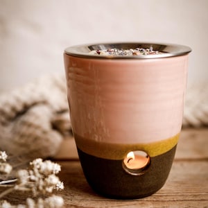 Könnte beinhalten: Ein rosa-brauner Keramik-Diffusor für ätherische Öle mit silbernem Rand. Der Diffusor hat eine kleine Öffnung mit einer brennenden Kerze im Inneren. Der Sockel ist dunkelbraun, mit einem gelben Band, das ihn vom rosa Oberteil trennt.