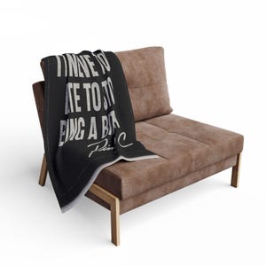 May include: A black throw blanket with white text reading "Time to be a bit..." is draped over a brown chair. The chair has a brown cushion and wooden legs. The blanket's text is in a bold, sans-serif font.