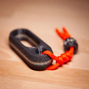 Könnte beinhalten: Ein dunkelgrauer, ovaler Schlüsselanhänger mit strukturierter Oberfläche und einer leuchtend orangefarbenen Kordel. Die Kordel ist an einer kleinen, dunkelgrauen Perle und einer Schlaufe befestigt. Der Schlüsselanhänger liegt auf einer Holzoberfläche.