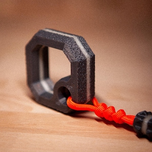 Può includere: Un moschettone nero ottagonale con superficie strutturata e striscia bianca. Una corda arancione è attaccata al moschettone. Il moschettone è su una superficie di legno. Questo articolo è adatto per attività all'aperto.