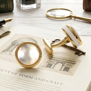 May include: A pair of round cufflinks featuring a polished gold-tone metal frame with a twisted rope border and a shimmering iridescent mother-of-pearl inlay. One cufflink is positioned upright, while the other lies flat, revealing a white side panel.