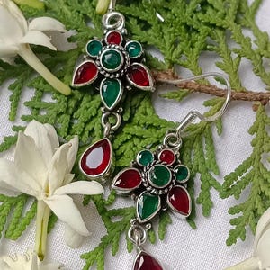 Pode incluir: Um par de brincos de prata com um design floral, com pedras preciosas vermelhas e verdes. Cada brinco tem um pendente de pedra preciosa vermelha em forma de gota. Os brincos são exibidos sobre uma cama de folhagem verde e flores brancas.