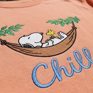 May include: Peach-colored sweatshirt featuring an embroidered Snoopy and Woodstock relaxing in a hammock under green leaves. The word "Chill" is embroidered in blue script below the image. A comfortable, casual top.