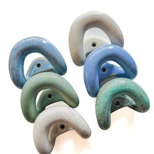 May include: A collection of eight U-shaped ceramic wall hooks arranged in two vertical columns. The hooks feature a smooth, glossy finish in various shades of blue, green, and grey, each with a single circular mounting hole at the base.