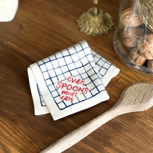 May include: A white ceramic spoon rest featuring a blue hand-painted grid pattern and the red text "even SPOONS must rest" written in the center. The object has a folded, rectangular shape with rounded corners and a smooth, glossy finish.