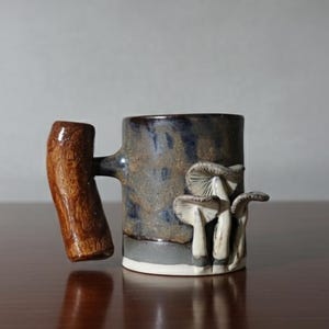 May include: A ceramic mug with a dark blue and brown glaze, featuring a wooden handle and a cluster of white mushroom sculptures on the side. The mug has a white base and is set on a dark wooden surface.