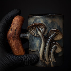 May include: A ceramic mug with a dark blue and gray glaze, featuring three-dimensional mushroom designs. The handle is a dark brown, wooden piece. The mug is held by a gloved hand against a black background.