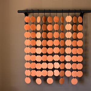 May include: A wall-mounted black metal rod supports multiple vertical strands of terracotta-colored circular discs. Each disc is connected to the next by small black rings, creating a grid-like hanging structure of uniform, flat, round clay-like pieces.