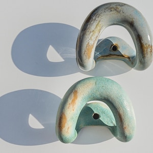 Könnte beinhalten: Zwei U-förmige Wandhaken aus Keramik mit glänzender Oberfläche. Der obere Haken zeigt eine Mischung aus hellblauen, grauen und braunen Tönen. Der untere Haken weist eine texturierte Mischung aus Türkis, Hellgrün und Beige auf, beide mit Loch.