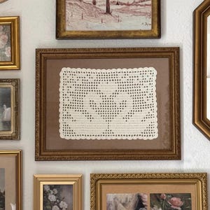 Puede incluir: Una colección de obras de arte enmarcadas y objetos decorativos en una pared blanca. Los marcos son de varios tamaños y formas, con tonos dorados y marrones. La obra de arte incluye una pintura de paisaje, estampados florales y un tapete de ganchillo enmarcado.