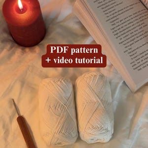 May include: A lit candle next to a book and two white yarn skeins. A crochet hook rests near the yarn. The image includes text that reads "PDF pattern + video tutorial". The scene is set on a soft, textured surface.