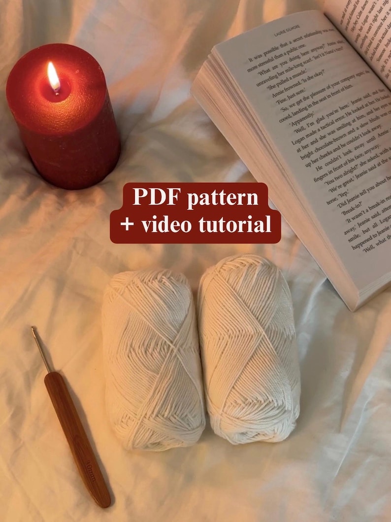 May include: A lit candle next to a book and two white yarn skeins. A crochet hook rests near the yarn. The image includes text that reads "PDF pattern + video tutorial". The scene is set on a soft, textured surface.