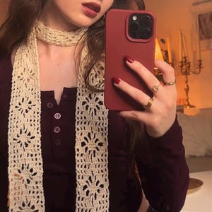 May include: A long, cream-colored crochet scarf featuring a repeating floral pattern with openwork details. The scarf is worn wrapped around the neck, paired with a dark maroon long-sleeved henley shirt that has a buttoned placket at the chest.