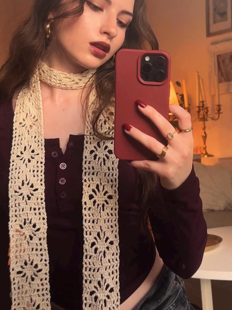May include: A long, cream-colored crochet scarf featuring a repeating floral pattern with openwork details. The scarf is worn wrapped around the neck, paired with a dark maroon long-sleeved henley shirt that has a buttoned placket at the chest.