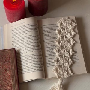 Op de afbeelding: Een handgemaakt, crèmekleurig gehaakt bladwijzer met bloemmotief en kwast, liggend op een open boek. Twee brandende rode kaarsen creëren een gezellige leessfeer.