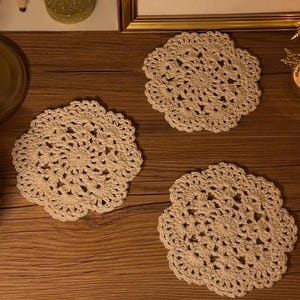 May include: Three cream-colored, handmade crochet doilies arranged on a wooden surface. Each doily has a floral design with a scalloped edge. The doilies are a neutral color, suitable for various home decor styles.